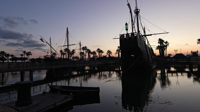 Cinematic wide shot of the historical replica ships. Pinta, Ni&ntilde;a and Santa Mar&iacute;a docked at Muelle de las Carabelas during a serene sunset. Located in La Rabida, Palos de la Frontera, Huelva, Spain.