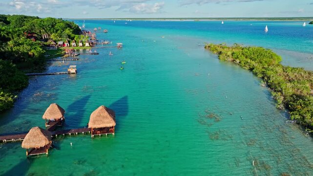 Aerial view of a thatched over-water palapa beside a turquoise channel in the Bacalar lagoon, Mexico, green mangrove islands dotting the clear water.