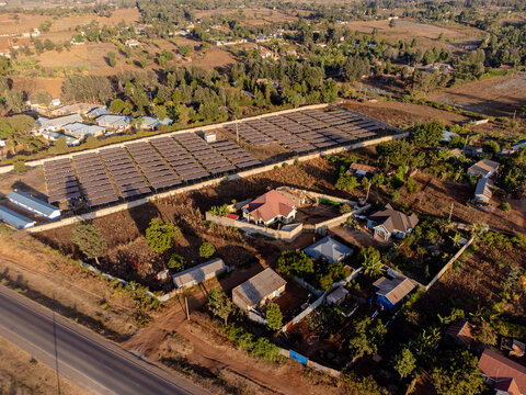 Aerial view of a solar farm with rows of photovoltaic panels, surrounded by residential houses, trees, and dry vegetation near a road in Loitokitok, Kajiado County, Kenya.