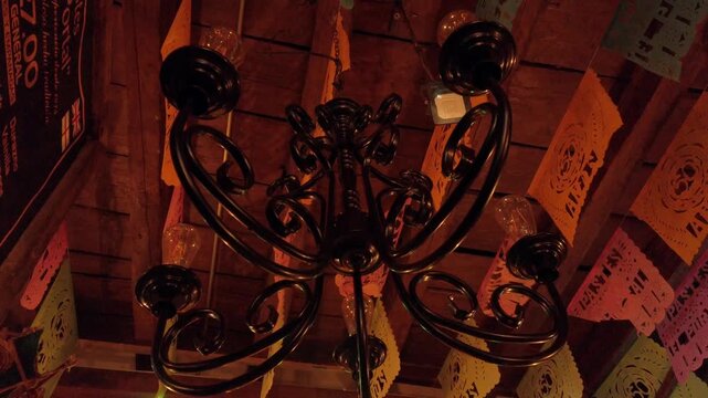 Low angle view of decorative wrought iron chandelier beneath wooden ceiling. Warm light illuminates colorful papel picado banners, highlighting traditional interior decor and detailed metalwork