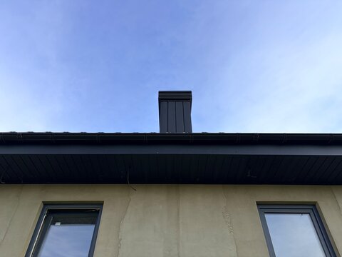 A modern house under construction showing black roof gutters, soffits, and unfinished plastered walls against a blue sky. Ongoing building process.
