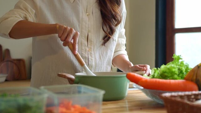 Woman Stirring Soup in Green Pot on Wooden Kitchen Counter with Fresh Vegetables