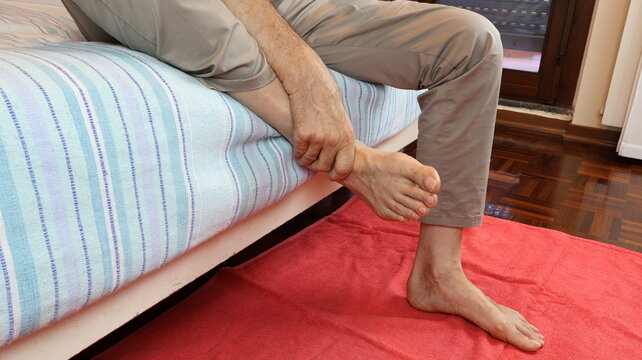 Elderly man painfully massages his ankle while sitting on bed, dressed in cotton trousers.