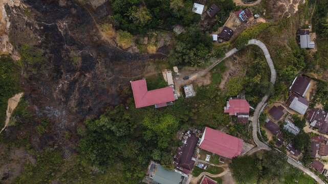 Aerial view of houses with red roofs nestled in a lush green landscape with a section of burnt vegetation and a winding road in Papua, Indonesia.