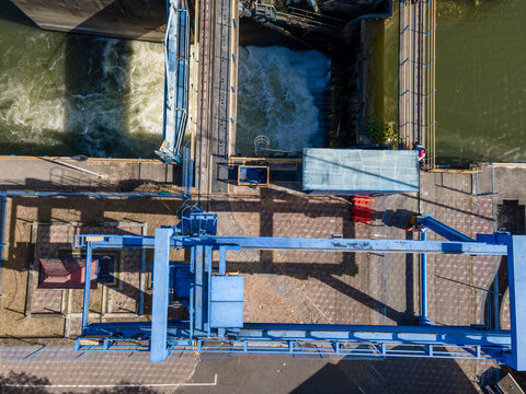 Aerial view of a water control structure with a blue metal gantry crane, rushing water through a sluice gate, and paved walkways in Banyumas Regency, Central Java, Indonesia.