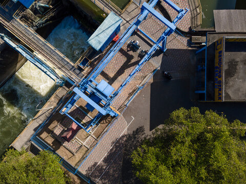 Aerial view of Bendung Gerak Serayu dam with blue metal sluice gate machinery, rushing water, paved walkways, and surrounding trees in Banyumas Regency, Central Java, Indonesia.