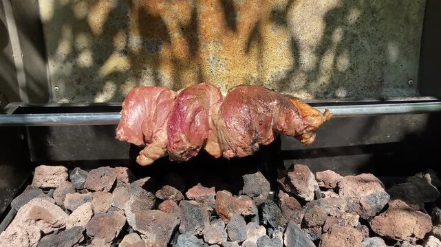 Close-up video frame of a Brazilian Picanha roast slowly rotating on a rotisserie above hot lava rocks in an outdoor barbecue grill. Juicy meat roasting over heat with smoke and natural sunlight, perf