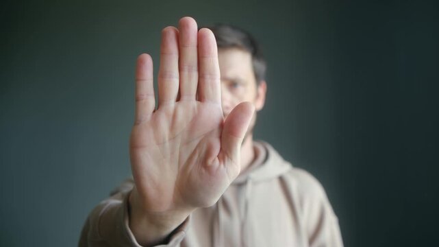Serious bearded caucasian man raising his hand to the camera making a stop gesture, expressing denial, rejection or prohibition against a plain studio background, focusing on the palm