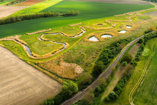 Aerial view of a meandering stream winding through lush green fields and agricultural land with a railway track running alongside in Cejkovice, South Moravian Region, Czechia.