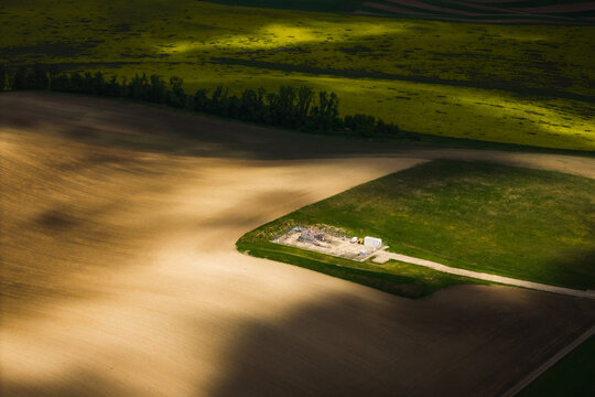 Aerial view of agricultural fields with a small industrial facility surrounded by green and brown soil under shifting cloud shadows in Brno, South Moravian Region, Czechia.