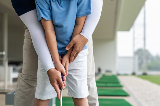Golf Coach guiding an Asian boy's hands into the correct club grip during a close-up junior golf lesson on a practice mat at a covered driving range