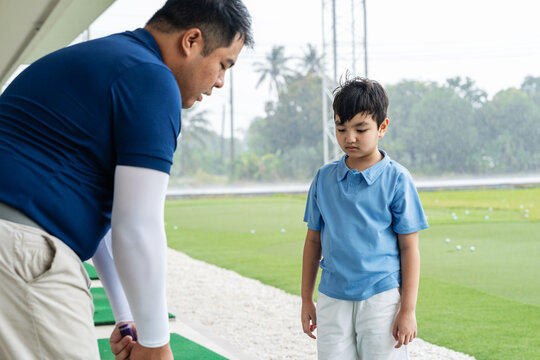 Junior Golf Lesson with an Asian boy watching a male coach demonstrate club placement and address position during beginner practice at a covered driving range