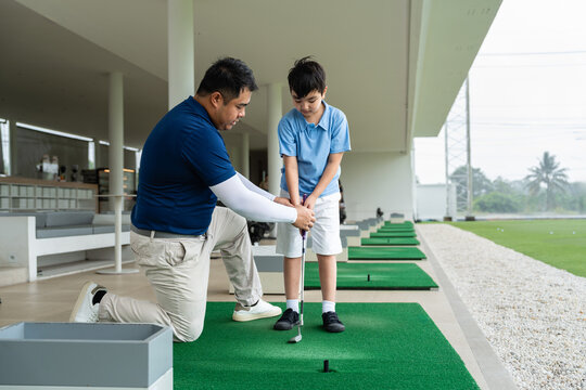 Junior Golf Lesson with a male golf coach kneeling to correct an Asian boy's grip and setup while practicing on a green mat at a covered driving range