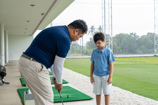 Junior Golf Lesson with a male coach demonstrating club setup and address technique while an Asian boy observes at a bright covered driving range