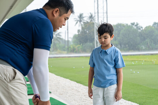 Golf Coach demonstrating proper club position to an Asian boy during a focused beginner golf lesson on a practice mat at a covered driving range