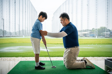 Fototapeta premium Junior Golf Lesson with a male golf coach kneeling to guide an Asian boy's arm position and stance while practicing with a club at a driving range