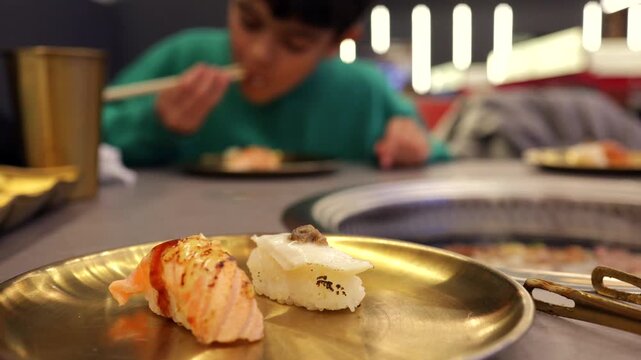 Close up of delicious seared salmon and scallop nigiri sushi with a young boy eating with chopsticks at a japanese restaurant in the blurred background