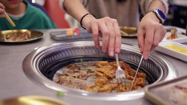 Woman's hands skillfully grilling marinated shrimp and thin slices of meat on an integrated tabletop korean barbecue grill at an authentic asian restaurant