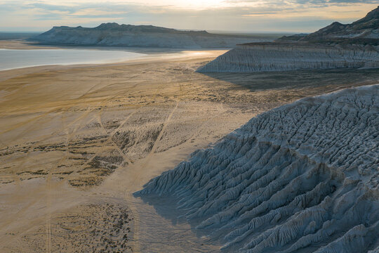 Aerial view of Tuzbair salt flats with white limestone cliffs, textured desert terrain, and tire tracks under soft golden hour light in Mangystau District, Mangystau Region, Kazakhstan.