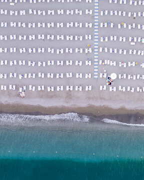 Aerial view of rows of white sun loungers arranged on a sandy beach with a blue walkway leading to the turquoise sea in Vietri sul Mare, Salerno, Campania, Italy.