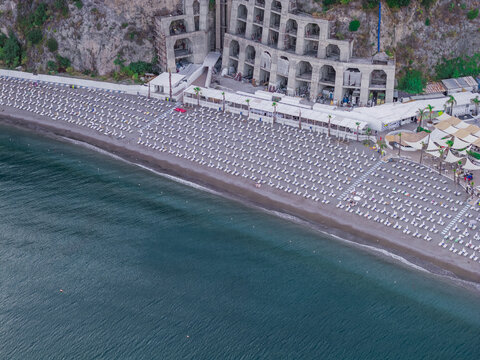 Aerial view of the beach with rows of sun loungers and umbrellas along the shoreline, adjacent to a cliffside structure in Vietri sul Mare, Salerno, Campania, Italy.