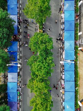 Aerial view of a bustling Car Free Day market with rows of blue tents, lush green trees in the median, and people walking between stalls in Samarinda, Kalimantan Timur, Indonesia.