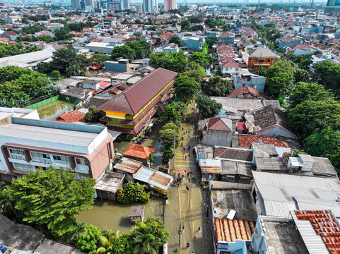 Aerial view of a flooded street surrounding the SDN Kembangan Utara 2 PT3 PG school building with residential rooftops and lush trees in Kecamatan Kembangan, Daerah Khusus Ibukota, Indonesia.