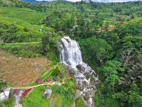 Aerial view of Curug Cikondang waterfall cascading over rocky cliffs, surrounded by lush green rice terraces, tropical vegetation, and small rural houses in Campaka, West Java, Indonesia.
