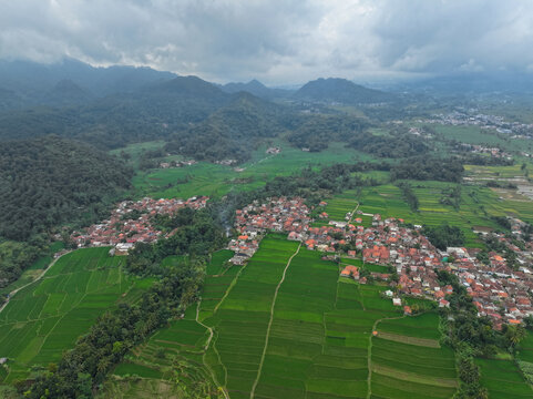 Aerial view of lush green rice terraces surrounding a village with terracotta rooftops nestled against rolling mountains under a cloudy sky in Campaka, West Java, Indonesia.