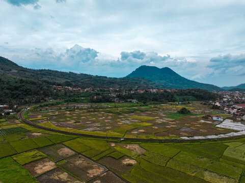 Aerial view of lush green rice fields and a winding railway track set against a backdrop of rolling mountains in Kadungora, West Java, Indonesia.
