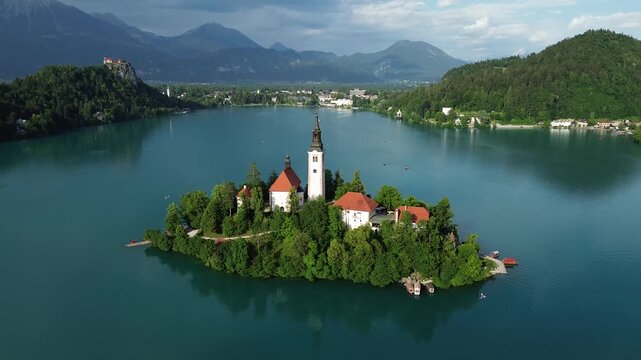Drone flight away from Bled Island on Lake Bled, Slovenia, featuring the Church of the Assumption, Bled Castle, turquoise water and the Julian Alps.