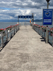 Fototapeta premium Lake geneva pier extending towards mountains under cloudy sky, decorated with vibrant flowers, showing a departure sign and clock indicating ferry travel