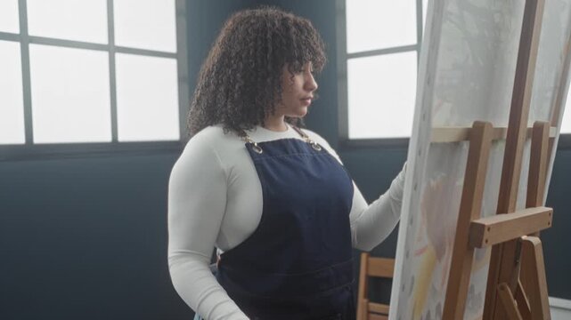 Woman painting on canvas in a studio, side profile wearing apron, arm reaching with paintbrush at easel near window light; quiet concentration.