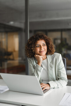 Vertical portrait of hispanic businesswoman resting chin on hand, using laptop at desk in office, thinking. Young indian business woman, project manager, considers negotiation offer, smiling.