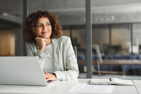 Chin resting on hand, indian businesswoman using laptop at desk in office, reviewing contract with smiling expression. Young hispanic business woman, auditor, gathers thoughts at corporate workplace.