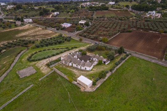 Aerial view of a traditional trullo complex with conical roofs surrounded by olive groves, stone walls, and agricultural fields in the rural landscape of Locorotondo, Apulia, Italy.