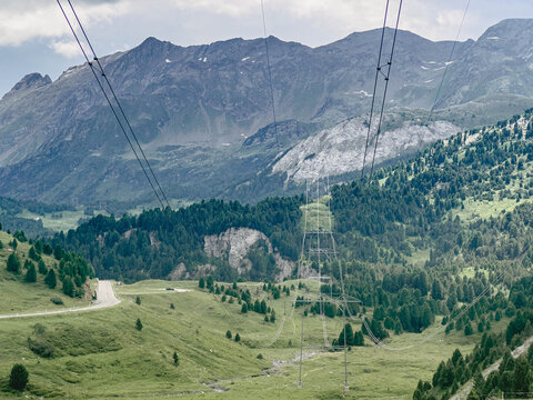 High-voltage power lines crossing a wide alpine valley surrounded by rugged mountain peaks and green meadows. Energy transmission infrastructure in a pristine natural environment.