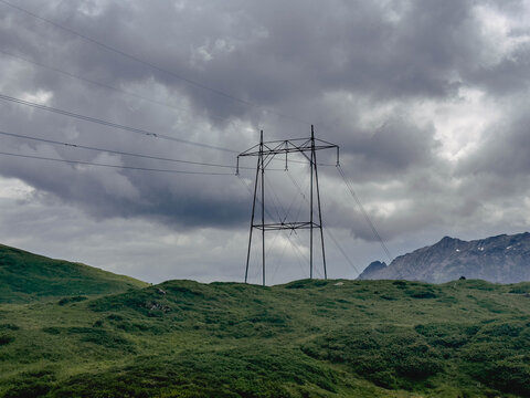 High-voltage transmission tower standing on a green alpine hillside beneath dramatic storm clouds. Renewable energy infrastructure in a remote mountain landscape with moody weather.