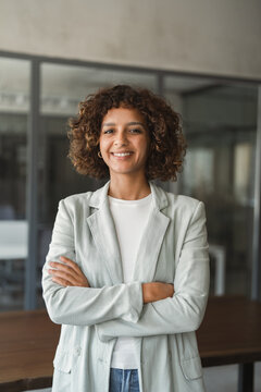 Vertical portrait of businesswoman standing in office with arms crossed, using confident posture, smiling toward camera. Young latina business woman poses as ceo, leader at workplace. Copy space