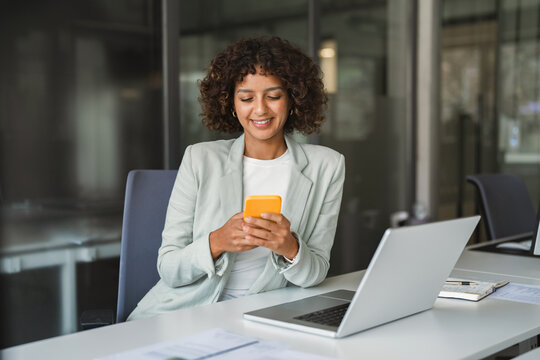 Sitting at desk in office beside open laptop, hispanic businesswoman using smartphone, smiling with focused expression. Young indian business woman, banking manager, works at corporate workplace.