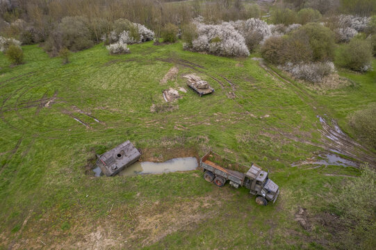 Aerial view of an abandoned army truck and a tank resting in a grassy field surrounded by trees and shrubs in Belgium.