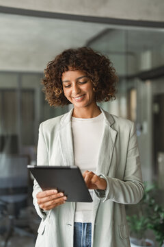 Vertical portrait of hispanic businesswoman standing in office, using tablet, smiling, working on sales report. Young indian business woman, marketing manager, reviews data at workplace. Copy space