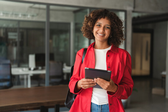 Portrait of curly haired hispanic woman wearing backpack, using tablet, smiling toward camera as student. Young latina girl captures casual, friendly lifestyle moment heading out for busy campus day.