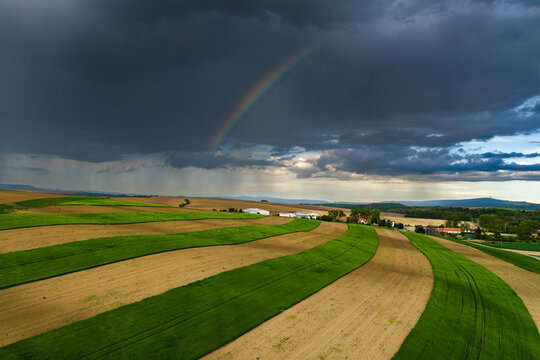 Aerial view of agricultural fields with alternating green and brown strips under a dark stormy sky with a rainbow in Nasedlovice, South Moravian Region, Czechia.