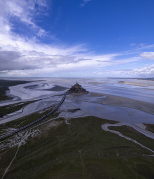 Aerial view of Mont Saint-Michel abbey surrounded by tidal flats and the causeway under a blue sky in Mont Saint-Michel, Normandy, France.