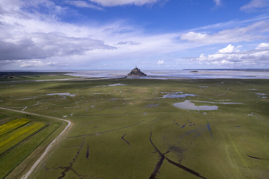 Aerial view of the historic abbey of Mont Saint-Michel surrounded by vast tidal flats and green salt marshes under a bright blue sky in Mont Saint-Michel, Normandy, France.
