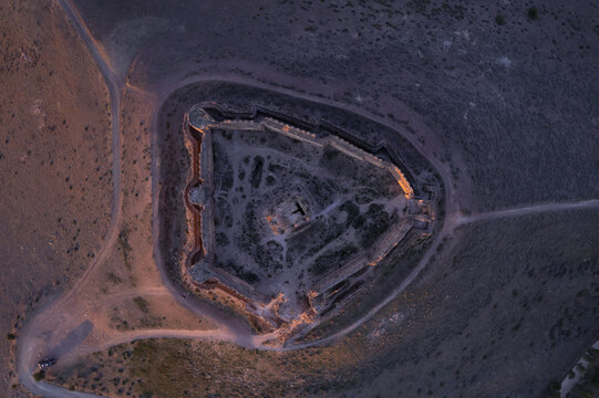 Aerial view of the ruins of Castle Almonacid de Toledo on a hilltop at sunset, with dirt paths around the historic stone fortifications in Almonacid de Toledo, Castilla-La Mancha, Spain.