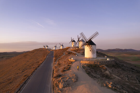 Aerial view of Molinos de Viento de Consuegra windmills on a hill at golden hour with a road and a person walking in Consuegra, Castilla-La Mancha, Spain.