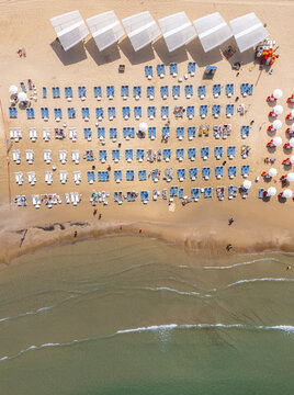 Aerial view of Herzliya Beach with rows of blue sun loungers and white sunshades arranged on the golden sand along the Mediterranean Sea in Herzliya, Tel Aviv District, Israel.