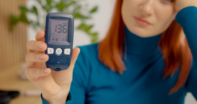 A woman holds a glucometer showing a reading of 136 mg dL. She appears concerned as she checks her blood sugar levels at home during the day.